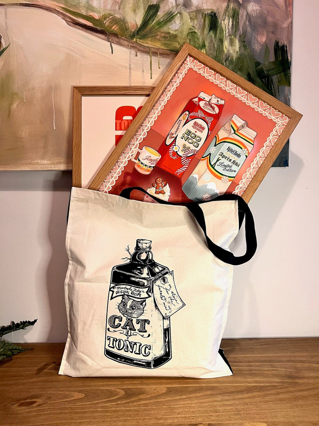 A white tote bag with a "Cat Tonic" design rests on wood, exuding festive gift shop vibes as it carries two colorful Kitsch Kanaveral x Hôtel Dodo Noggin’ The Nog Giclée Art Prints by Hôtel Dodo, both featuring illustrated bottles.