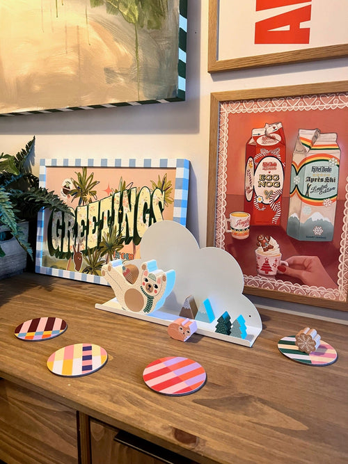 At Hôtel Dodo, a wooden shelf displays plants, striped coasters, a bear decoration, and the “Noggin’ The Nog” Giclée Art Print by Kitsch Kanaveral x Hôtel Dodo—perfect for a festive gift shop vibe.