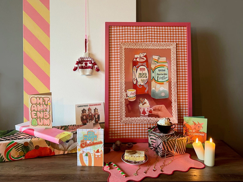 A festive display with Hôtel Dodo's Kitsch Kanaveral x Hôtel Dodo Noggin’ The Nog Giclée Art Print (milk and egg nog cartons), holiday cards, gifts, candles, a Christmas mug, cake plate, and seasonal decor—perfect for any gift shop.