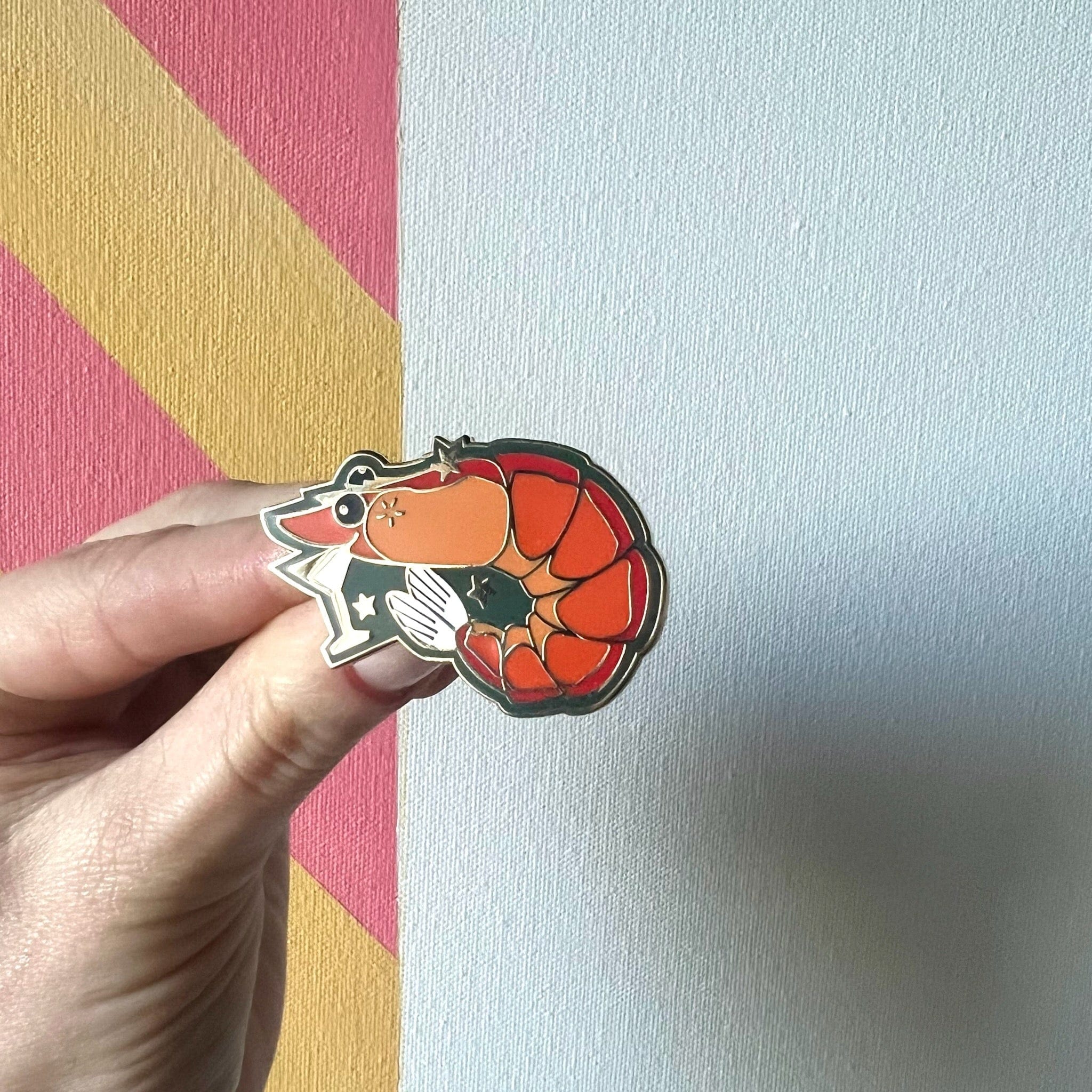A hand holds the Prawn Cocktail Pin by Pins by diedododa, featuring an orange and black cat curled into a prawn shape. The background is a white wall with pink and yellow diagonal stripes, reminiscent of lively cocktail party decor.