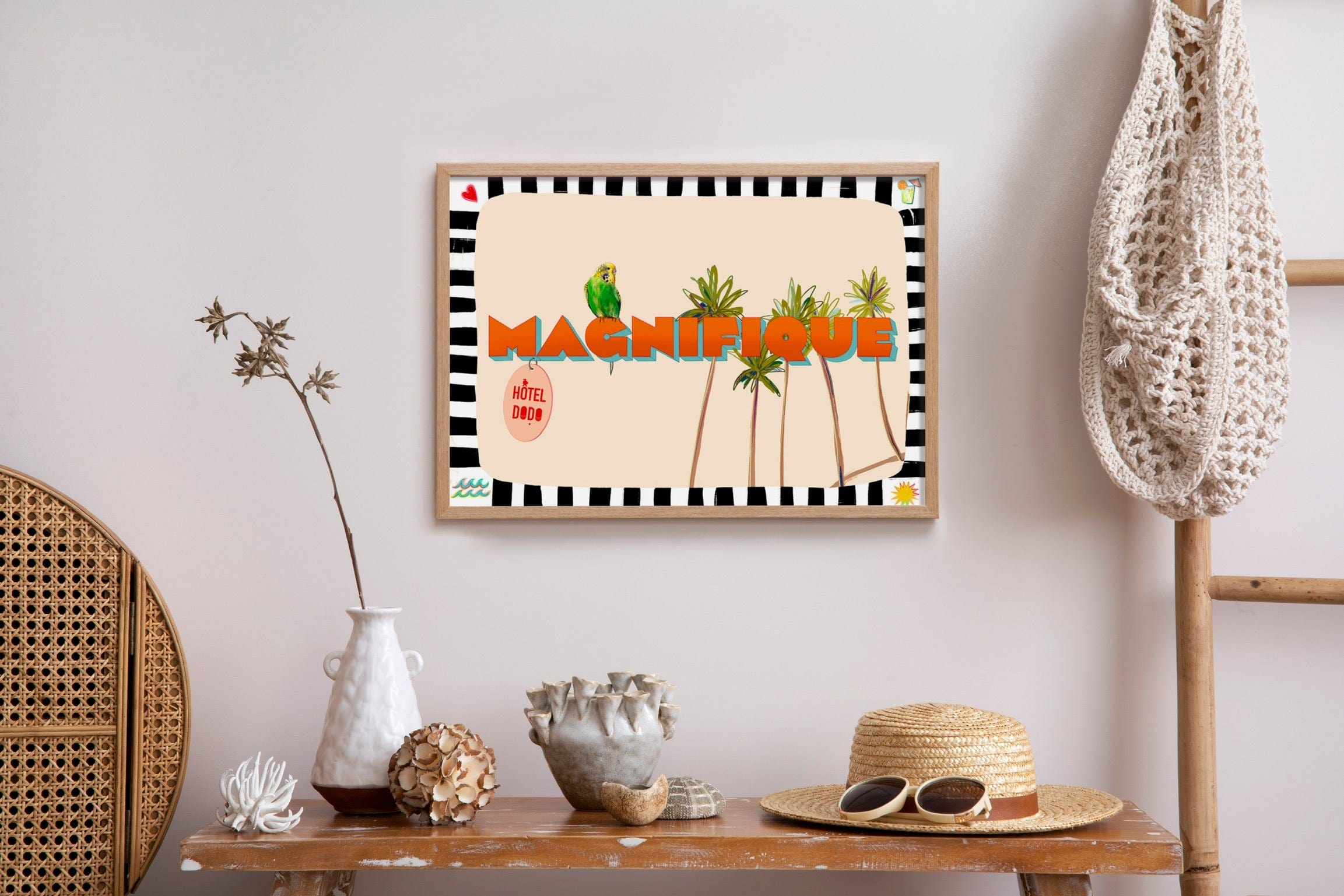 The Shromps "Magnifique" Matte Art Print, featuring palm trees and a parrot, brings Hôtel Dodo Resort vibes above a wooden table with a vase, shells, straw hat, and a crochet bag hanging nearby.
