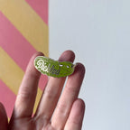 A hand displays the Pickle Pin by Pins by diedododa, featuring a tiny pickled pickle against a vibrant pink and yellow striped background with "Pickled" proudly shown.