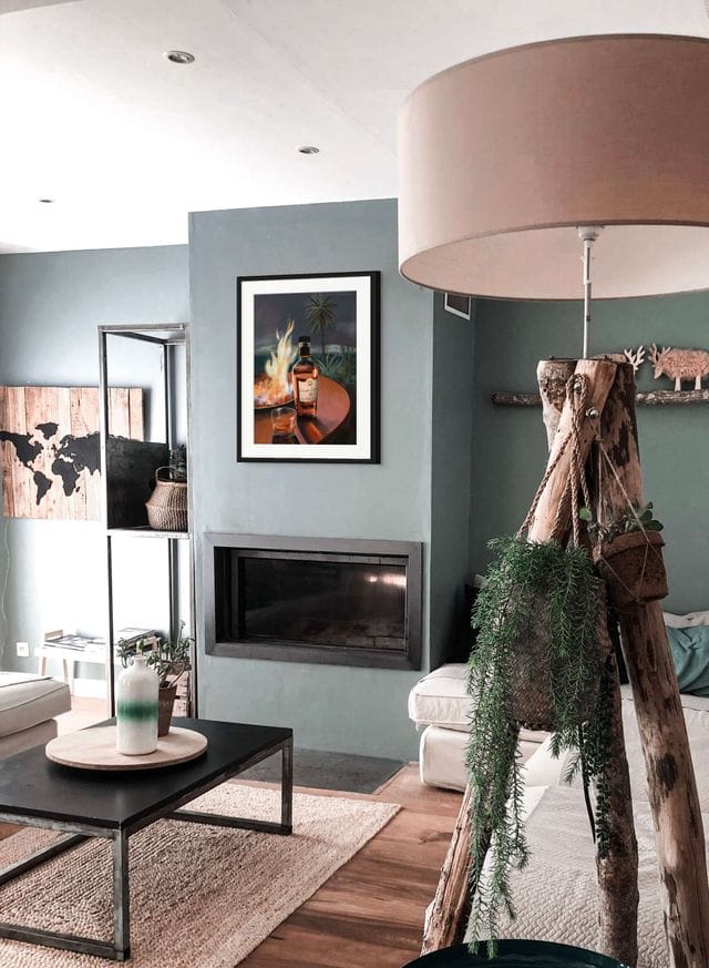 Modern living room with a fireplace, neutral furniture, curated coffee table settings, a large floor lamp, and green plants on a wooden stand. The Hôtel Dodo Scottish Riviera Giclée Art Print and candles add charm to this inviting space.