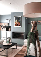 Modern living room with a fireplace, neutral furniture, curated coffee table settings, a large floor lamp, and green plants on a wooden stand. The Hôtel Dodo Scottish Riviera Giclée Art Print and candles add charm to this inviting space.