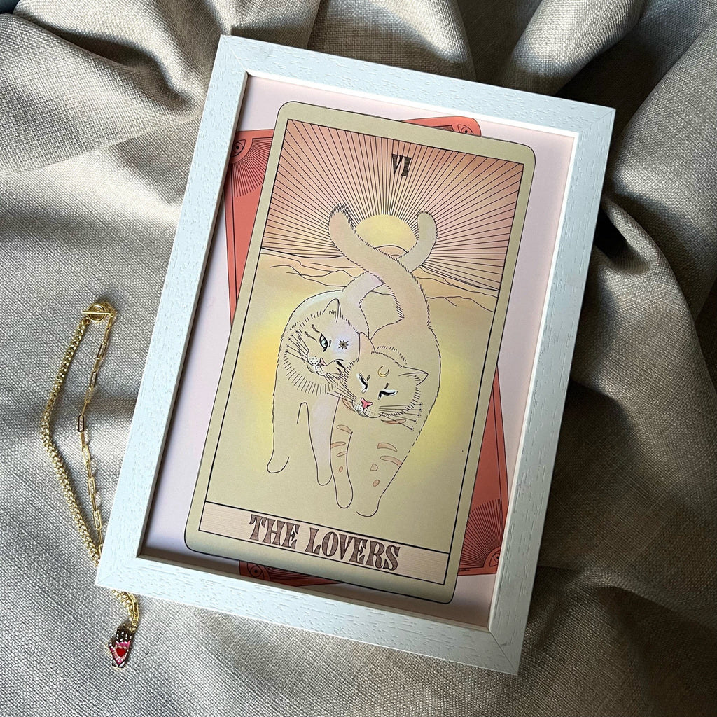 The Lovers Matte Art Print by Tarot Cats features two cats with intertwined tails and is shown on grey fabric next to a red pendant necklace.